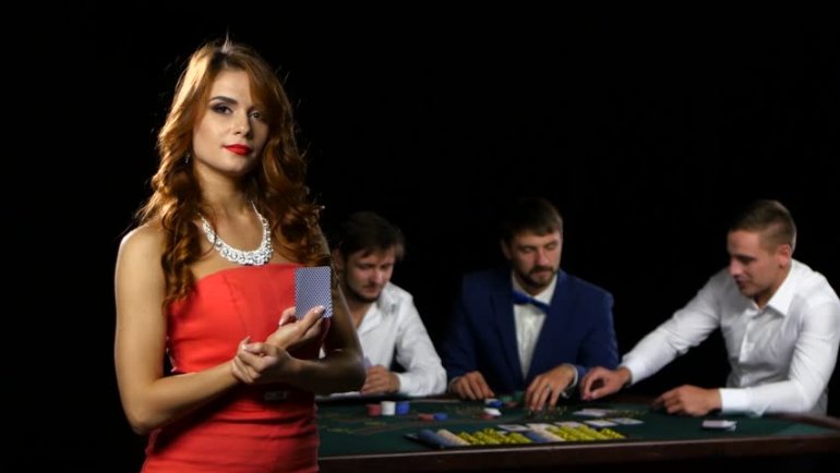 Рыжеволосая красотка в красном обтягивающем платье стоит с картой в руках, а на заднем фоне трое мужчин играют в покер