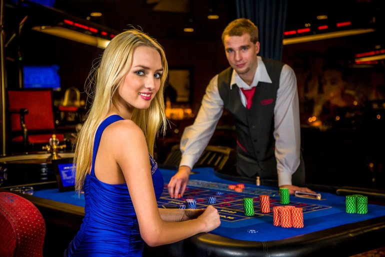 Привлекательная блондинка в обтягивающем ярко-синем платье сидит за игорным столом для рулетки, а молодой крупье готовится принять ставку