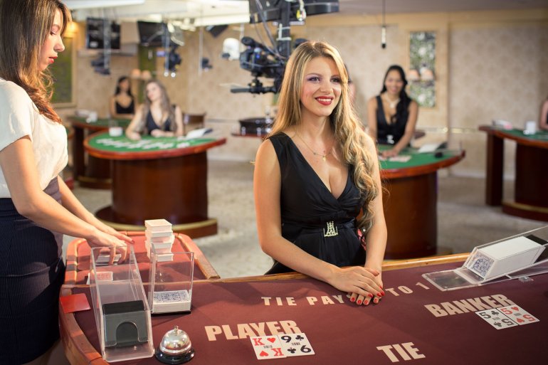 Стройная блондинка в черном платье с откровенным декольте ждет игроков у стола для баккара