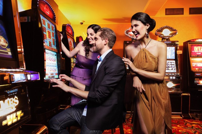Мужчина в окружении красиво одетых женщин сидит за игровым автоматом