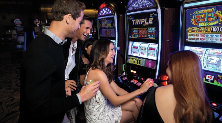 Девушки в хорошем настроении играют в игровые автоматы, а мужчины за их спинами наблюдают за экраном