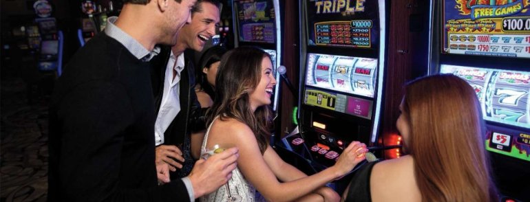 Молодые красивые девушки играют на автоматах в казино, а их парни наблюдают за игрой, попивая коктейли