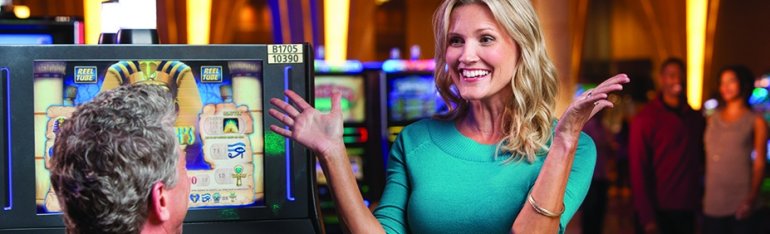 Женщина не сдерживает своего восторга от успеха в казино, а ее спутник восторженно смотрит на нее, сидя за игровым автоматом