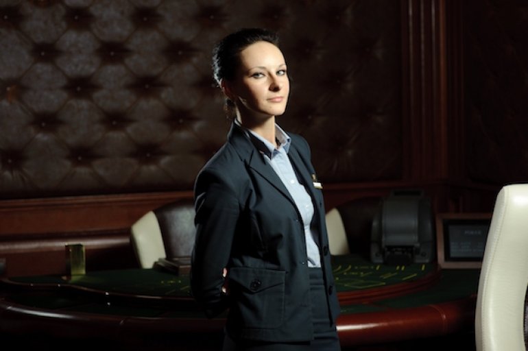Строгая женщина питбосс в деловом костюме стоит в зале казино рядом со столом для игры в блэкджек