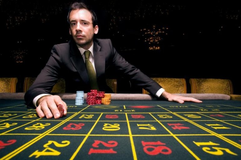 Задумчивый мужчина в строгом костюме с фишкой в руках сидит за столом для игры в рулетку
