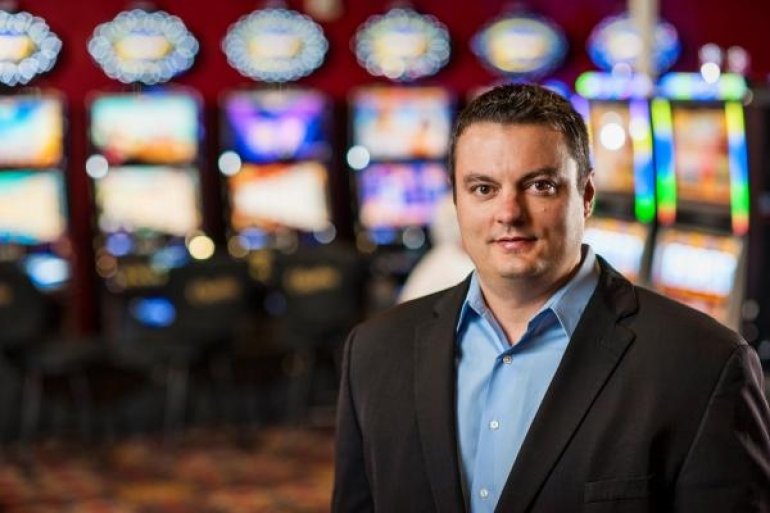 Питбосс в деловом костюме стоит в зале казино на фоне ряда игровых автоматов