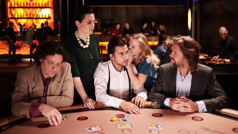 Привлекательная блондинка нашептывает на ухо своему партнеру, который сидит за столом для покера с другими игроками