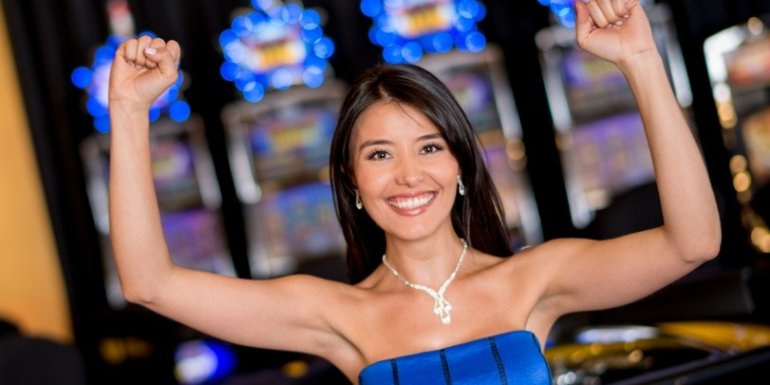 Симпатичная девушка в синем платье подняла руки вверх, радуясь своему выигрышу в казино на игровых автоматах